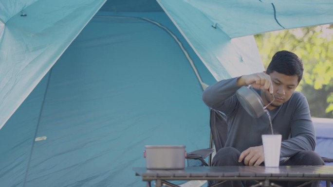 早上在露营地，亚洲男旅行者坐在野营椅上喝咖啡。