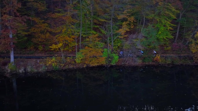早晨从空中俯瞰一个黑暗的、反光的湖，岸上有五颜六色的秋天的树木。摄影车在平静的水面上跟随一小群公路自