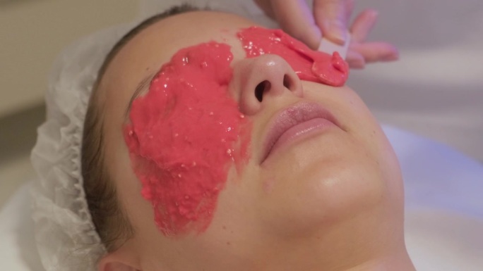 美容师用抹刀在女士脸上涂抹海藻酸盐面膜，特写。面部皮肤护理，美容院抗衰老程序。