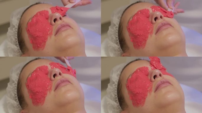 美容师用抹刀在女士脸上涂抹海藻酸盐面膜，特写。面部皮肤护理，美容院抗衰老程序。