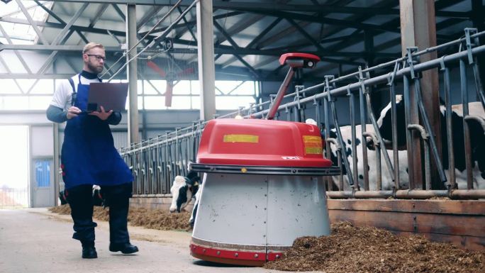 一位拿着笔记本电脑的农场专家正在跟踪一台自动推送饲料的机器