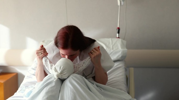慈爱的母亲坐在医院的病床上，把她的孩子抱在腿上，和她开玩笑地亲吻和说话
