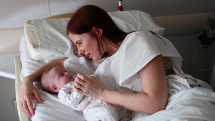 美丽的母亲躺在医院的病床上和她的女婴开玩笑地说话