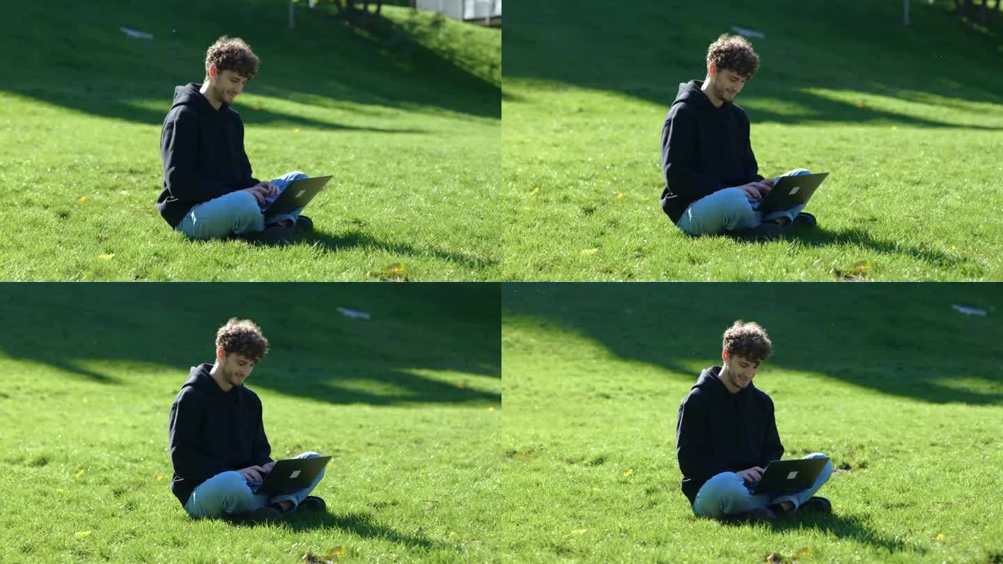 微笑的年轻人坐在公园的草地上，一边用笔记本电脑打字一边工作。秋天的季节。教育，工作理念。实时概念。
