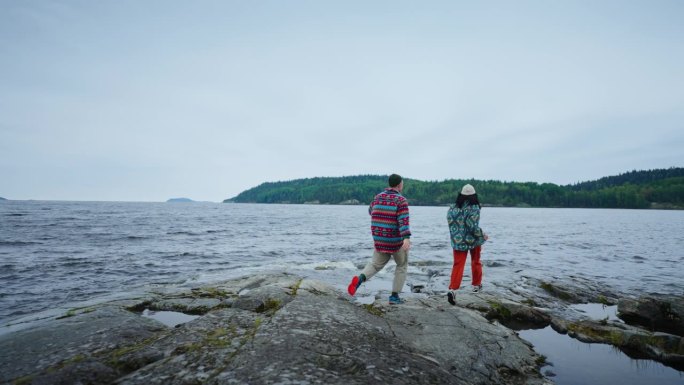 在斯堪的纳维亚美丽湖泊的石岸上，无忧无虑的情侣手牵着手奔跑，后视图