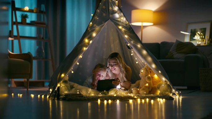 晚上，妈妈和孩子在客厅的帐篷里过夜，在家里用平板电脑阅读和放松。孩子们在黑暗的房子里露营，在公寓里和