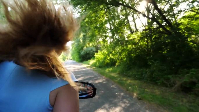 年轻的金发女孩把手伸出车窗，开着车，享受着旅行。一个无忧无虑的女人从车里探出身子，伸出手臂，骑着车穿
