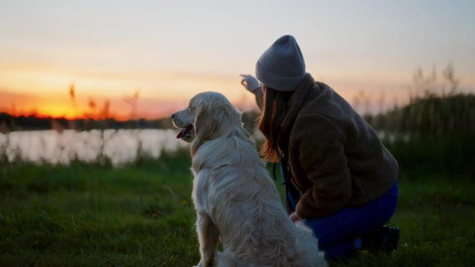一个女人和一只金毛猎犬坐在湖边的草地上看日落