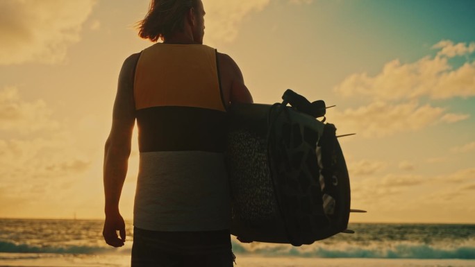 拿着冲浪板的男性冲浪者在日落的海滩上观看海浪。男性冲浪者站在冲浪板和观看海浪从阳光明媚的日落海滩，享