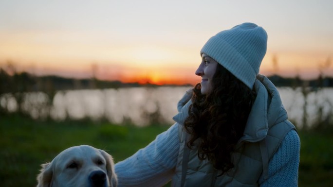 日落时分，一名妇女和一只金毛猎犬在湖边的草地上休息。抚摸和拥抱狗