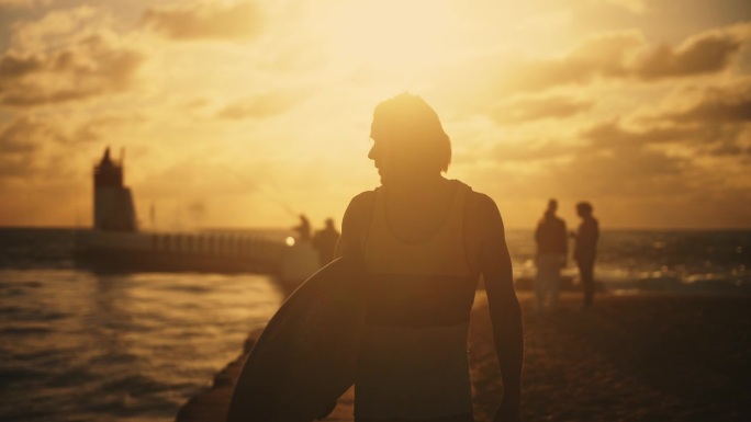 夕阳下夏日沙滩上，男性冲浪者手持冲浪板的剪影