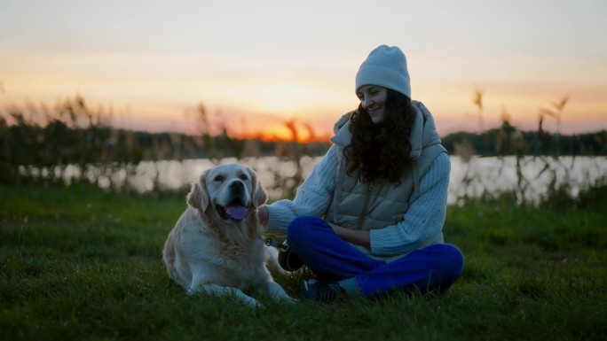 黄昏时分，一个女人和一只金毛猎犬坐在湖边的草地上。抚摸狗