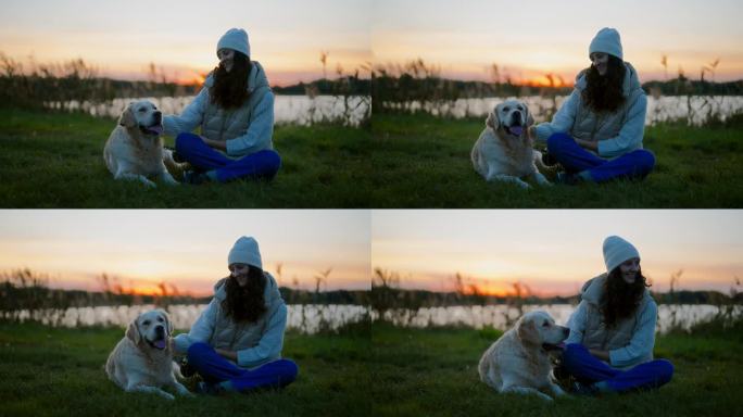 黄昏时分，一个女人和一只金毛猎犬坐在湖边的草地上。抚摸狗