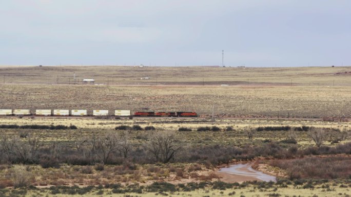 一列遥远的货运列车进入画面左侧，穿过亚利桑那州石化森林国家公园内的沙漠景观。前景是一条干涸的小河。