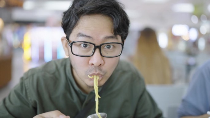 一名亚洲男子在一家食品中心与同事共进晚餐时，用勺子舀起鸡蛋面，向镜头展示，然后看着镜头，开心地吃着，
