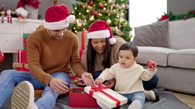 一对夫妇和儿子坐在家里圣诞树旁的地板上拆礼物