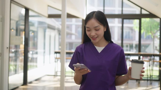 快乐的亚洲女护士，穿着紫色的手术服，拿着咖啡杯，从自助餐厅走回来