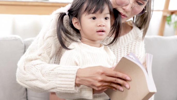 亚洲漂亮的孕妇妈妈在闲暇时间和大女儿一起在客厅看书，妈妈教宝宝读书，给小孩子讲故事。家庭幸福观