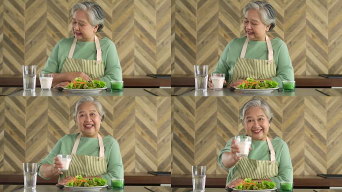 亚洲老年妇女早上在家里的厨房里喝着一杯牛奶，脸上挂着幸福的笑容。退休人员喝牛奶使骨骼强壮。预防骨质疏