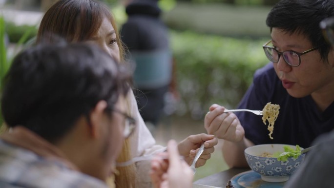 图为一名兴高采烈的女大学生在一所大学自助餐厅的户外，一边用叉子吃面一边和同学们讨论的特写。