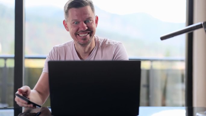 男人坐在桌子旁，在智能手机和笔记本电脑上阅读电子邮件，做出“是”的手势，感到快乐