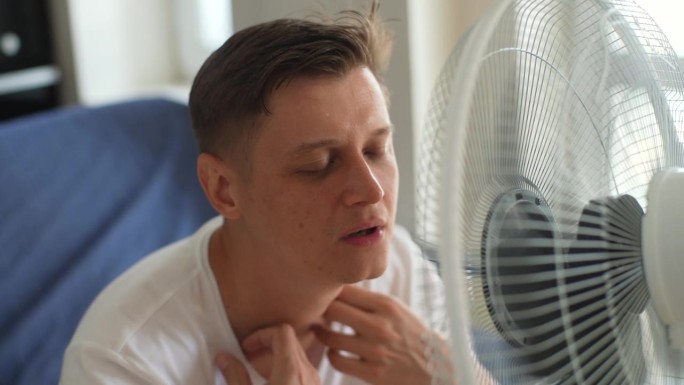 一名饱受酷暑折磨的疲惫男子试图在家中用电扇降温。大汗淋漓的男性坐在沙发上，用风扇冷却器散热