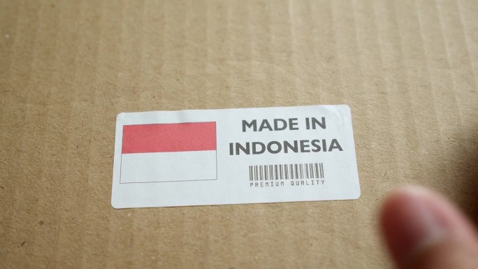 用手将印度尼西亚制造的旗帜标签贴在带有产品优质条形码的运输箱上。制造和交付。产品出厂进出口。