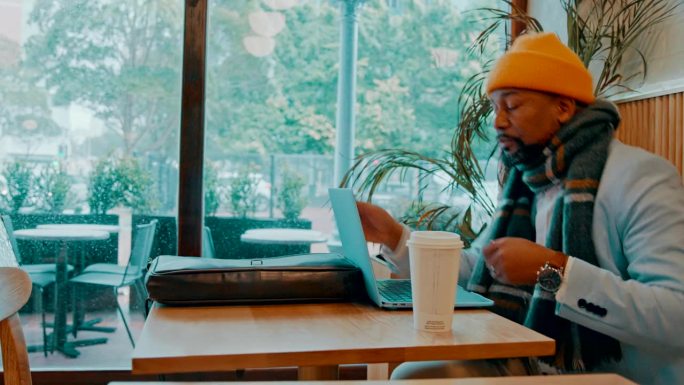 咖啡馆，咖啡和一个黑人男子用笔记本电脑远程工作，检查电子邮件或连接。冬天，餐馆和一个非洲企业家或成熟