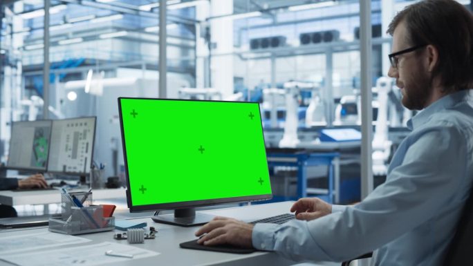 在工厂办公室里，机器人工程师在一台带有绿屏Chromakey模拟显示器的台式电脑上工作。具有自动装配