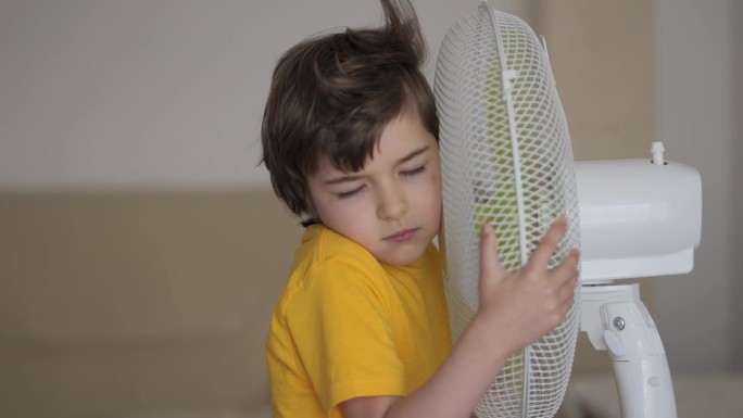 孩子在家里享受电风扇吹来的凉风。男孩在通风机前遭受高温，用电风扇冷却器给自己降温。炎热气候变化。