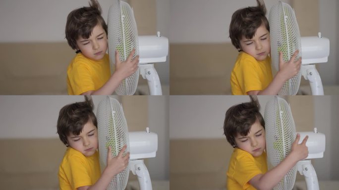 孩子在家里享受电风扇吹来的凉风。男孩在通风机前遭受高温，用电风扇冷却器给自己降温。炎热气候变化。