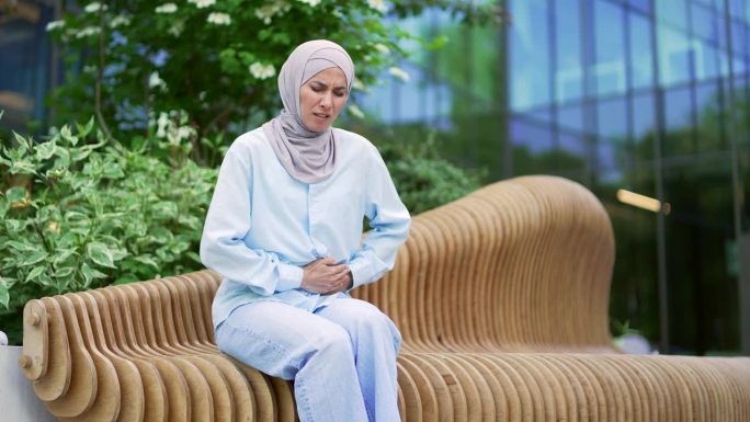 生病的年轻穆斯林女员工戴着头巾，胃痛地坐在办公大楼附近的长凳上