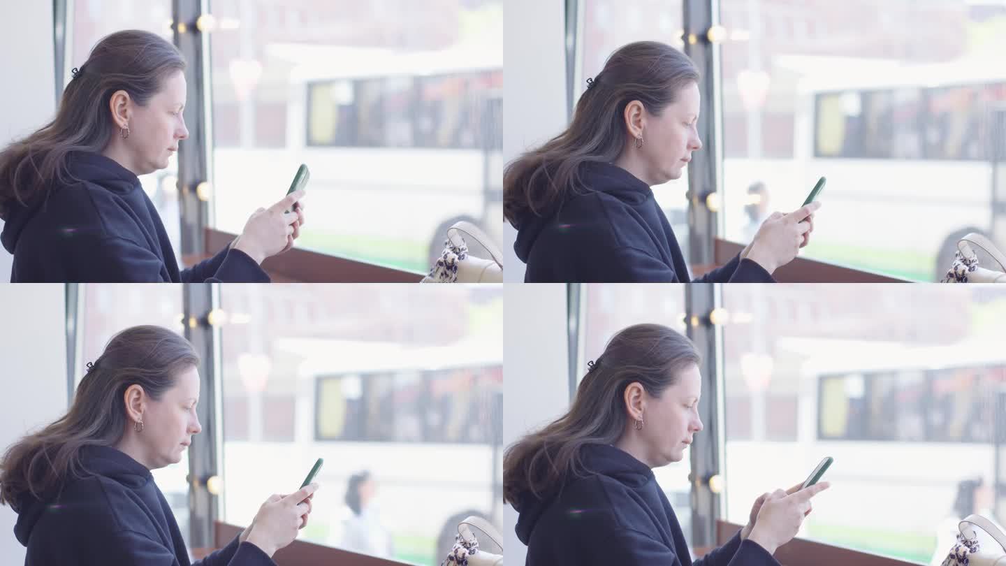 穿着深色衣服的女子坐在咖啡馆靠窗的地方，手里拿着智能手机在打字