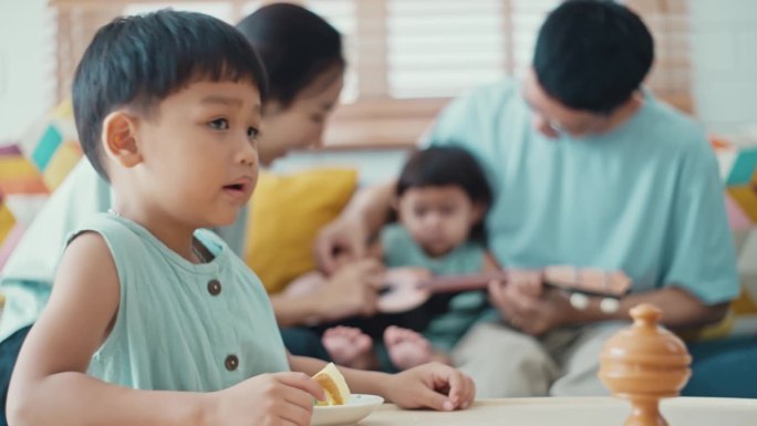 一个小男孩正在家里吃蛋糕，而他的父母和妹妹正在弹吉他