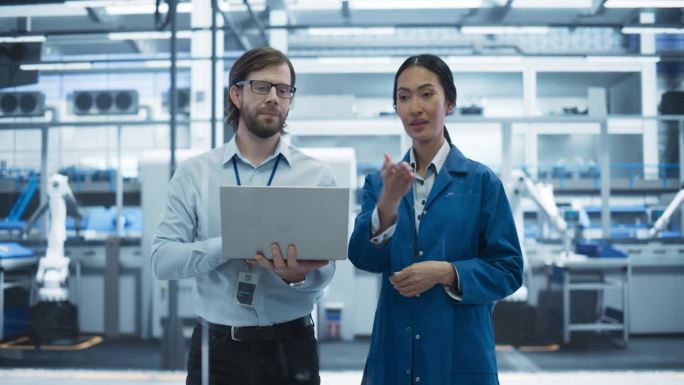 一名白人男子和一名亚洲女子在工业设施内交谈。先进的工厂员工协同工作，使用笔记本电脑监督工厂的工作过程