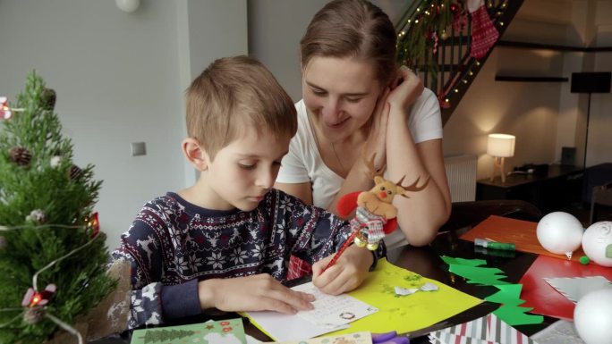 妈妈开心地微笑着看着儿子给圣诞老人写信。寒假，家人团聚，孩子和父母一起庆祝