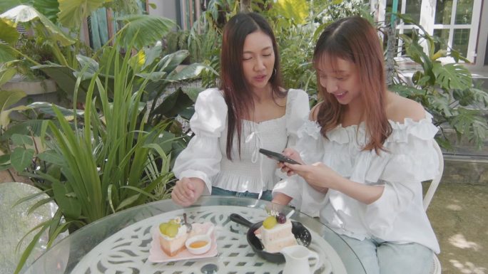 数码时代的两个亚洲美女的4K慢镜头，展现了在户外咖啡馆里通过智能手机保持联系，聊天的快乐和团聚。