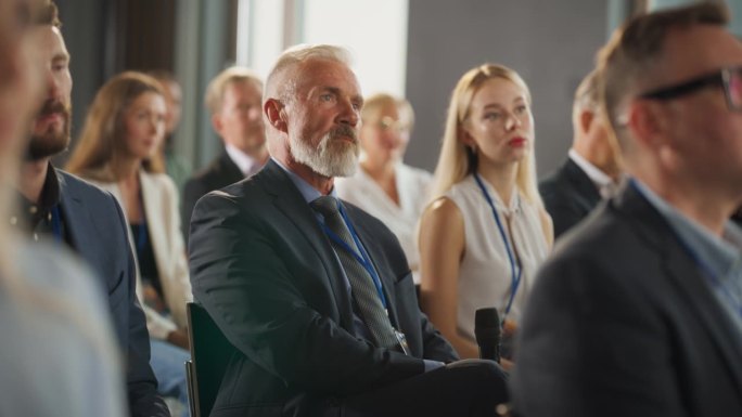 在国际科技会议上，资深男性投资者坐在人群中，用麦克风提问。一名白人男子正在听关于新公司服务的主题演讲