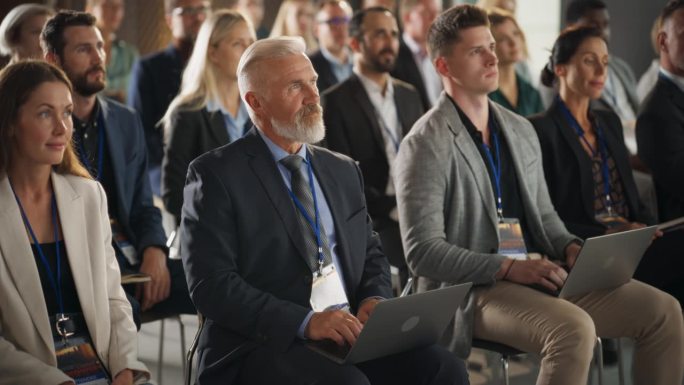 在商业论坛上，一位年长的白人男子坐在拥挤的听众中。公司代表使用笔记本电脑。成功的男性CEO参加商务会