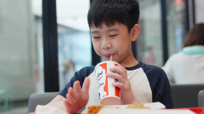 亚洲男孩在热闹的快餐店的快乐用餐体验。孩子坐在一张光线充足的桌子旁，脸上挂着欢快的微笑，津津有味地品