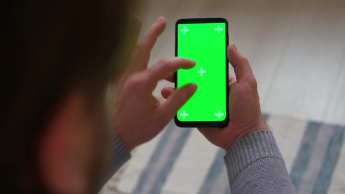 年轻人坐在家里，手里拿着智能手机的绿色模型屏幕。男性使用chromakey手机。垂直模式。触摸、滑动
