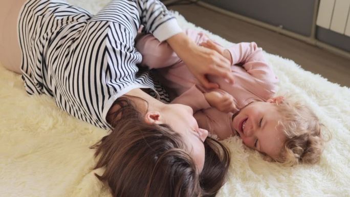 非常快乐乐观的年轻棕色头发的女人和她的小婴儿在家里躺在床上挠孩子，开心地笑着表达积极的情绪和幸福。