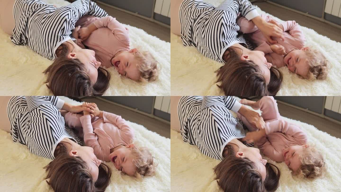 非常快乐乐观的年轻棕色头发的女人和她的小婴儿在家里躺在床上挠孩子，开心地笑着表达积极的情绪和幸福。