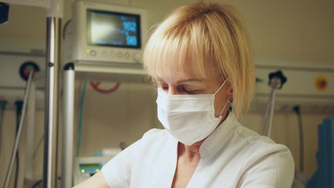 一名戴着防护口罩的金发女医生正在照顾一个生病的婴儿，为恒温箱打开或穿衣。新生儿重症监护。