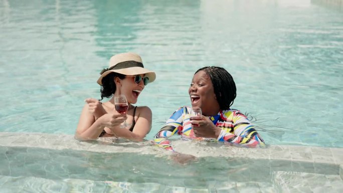 非洲女人和西班牙女人，两个亲密的朋友，多民族的人，在泳池里喝着酒，开心地聊天。在炎热的晴天里，在游泳
