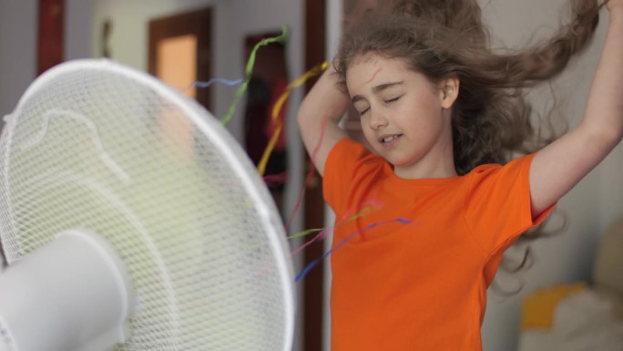 酷暑的孩子们享受着吹着风扇的清凉微风。大热天在家里的客厅里。炎炎夏日，孩子在家享受电风扇带来的凉风。