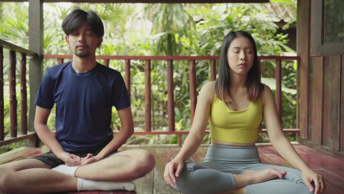 年轻的亚洲夫妇穿着运动服正在家里冥想，练习瑜伽。一对亚洲异性恋夫妇在家里的木阳台上一起冥想。在家运动
