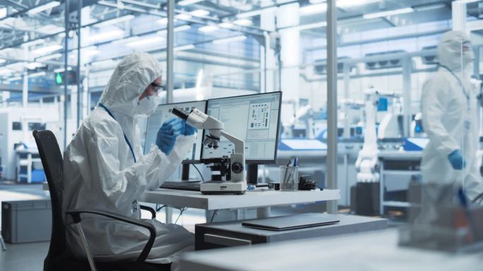 现代工厂:工业工程师穿着干净的无菌工作服在台式电脑前工作。科学家使用显微镜，为高科技医学疫苗和基因研