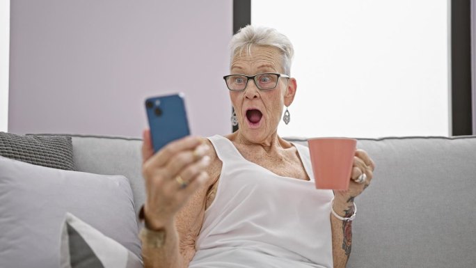 哇!惊讶的白发老人，惊讶地张大嘴巴，一边在家放松一边用智能手机喝咖啡