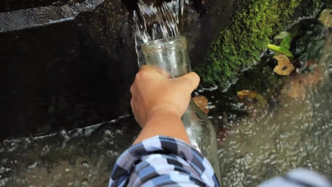 一个人把泉水装进透明的瓶子里的特写，这是一种美丽的天然清洁饮用水来源。一只手拿着一个瓶子，放在一股凉